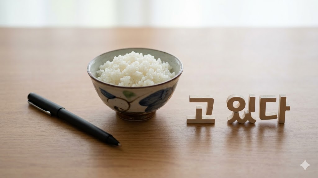 木のテーブルに置かれた黒ペン、ご飯茶碗、そしてHangeulの「고 있다」の文字ブロック。動詞の語幹にそのままつけるだけのシンプルな接続イメージ。