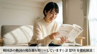 リビングで韓国語のテキストを手に笑顔を見せる日本人女性。画像下部に「韓国語の動詞の現在進行形：〜しています（-고 있다）を徹底解説！」という記事タイトルテキストが重なっている。